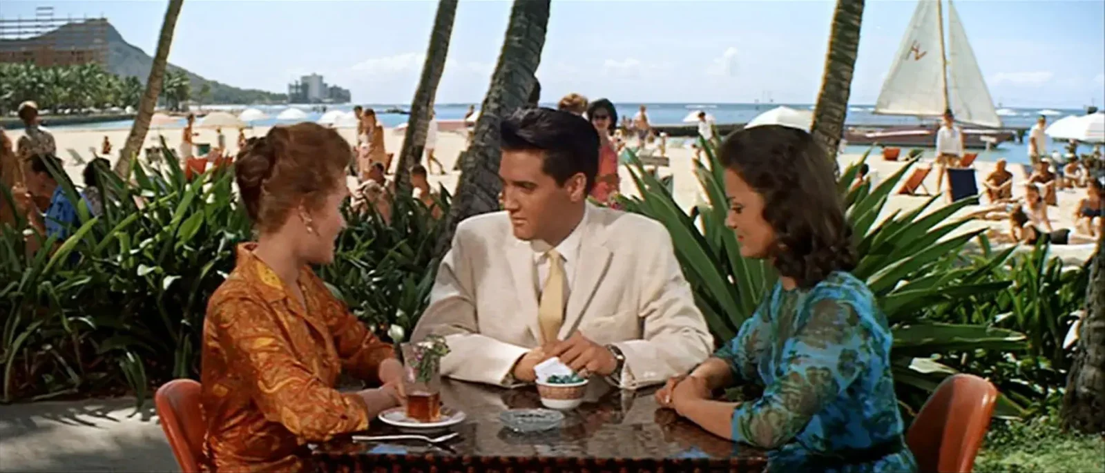 Elvis Presley seated with two women at an outdoor table beside Waikīkī Beach in Blue Hawaii